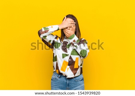 latin american woman covering face with both hands saying no to the camera! refusing pictures or forbidding photos isolated against yellow wall