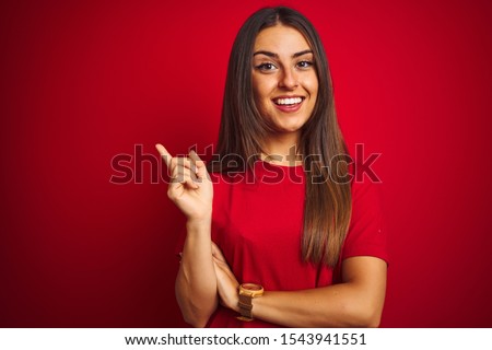 Young beautiful woman wearing t-shirt standing over isolated red background with a big smile on face, pointing with hand and finger to the side looking at the camera.