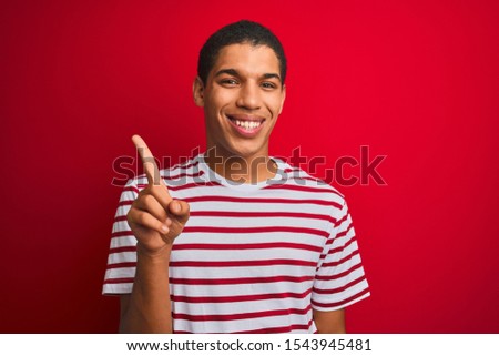 Young handsome arab man wearing striped t-shirt over isolated red background showing and pointing up with finger number one while smiling confident and happy.