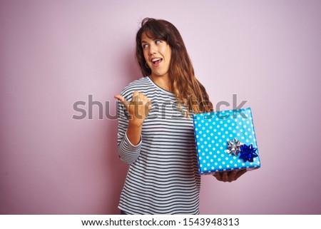 Young beautiful woman holding birthday gift over pink isolated background pointing and showing with thumb up to the side with happy face smiling