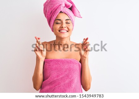 Young beautiful woman wearing shower towel after bath over isolated white background gesturing finger crossed smiling with hope and eyes closed. Luck and superstitious concept.
