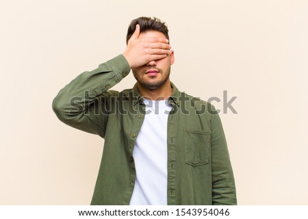 young hispanic man covering eyes with one hand feeling scared or anxious, wondering or blindly waiting for a surprise against isolated background