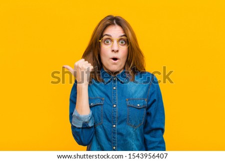 young pretty woman looking astonished in disbelief, pointing at object on the side and saying wow, unbelievable against yellow background