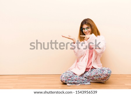 young woman wearing pajamas sitting at home smiling cheerfully and pointing to copy space on palm on the side, showing or advertising an object