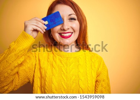 Young beautiful redhead woman holding credit card over yellow isolated background with a happy face standing and smiling with a confident smile showing teeth