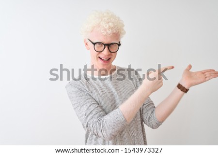 Young albino blond man wearing striped t-shirt and glasses over isolated white background amazed and smiling to the camera while presenting with hand and pointing with finger.