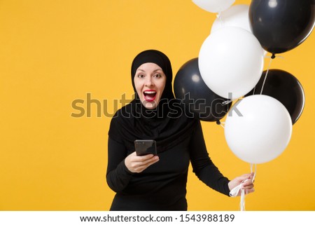 Arabian muslim woman in hijab celebrating hold cellphone black white air balloons isolated on yellow background studio portrait. Birthday holiday people religious lifestyle concept. Mockup copy space