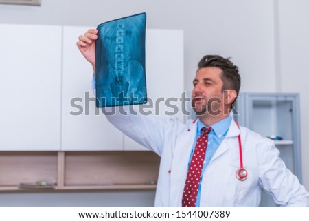 Male Doctor in his mid 40's reading an x-ray image ( radiography ) from a patient hip region (coxa) while standing in a hospital radiology department.
