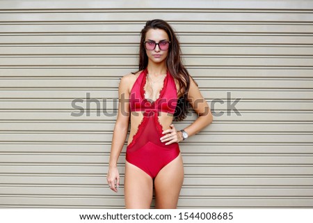 Elegant young brunette woman in bikini and sunglasses posing against grey wall