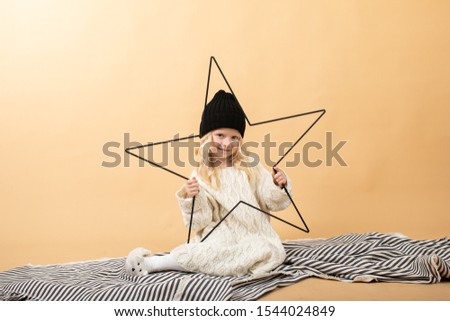 A little girl in a white knitted dress and a black hat sits on a plaid on a yellow background. Girl holding a big star