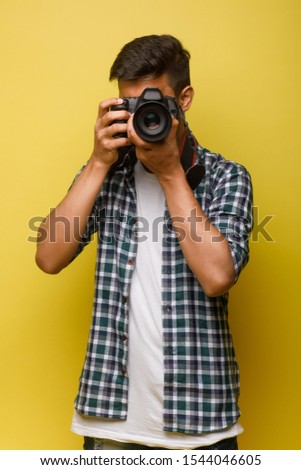 Handsome and confident indian man photographer with a large professional camera taking pictures photo shooting on the on the yellow background. Lifestyle.