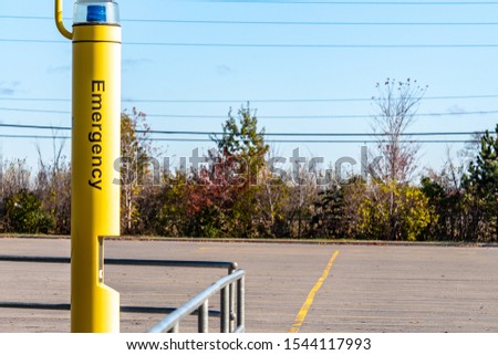 A yellow pole standing in a public parking lot features a blue light and an intercom for requesting help. The word "Emergency" appears on the side of the lot's safety feature.