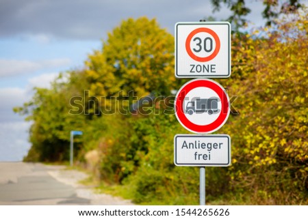 Three signs on a street. Ban on trucks. Speed limit in the zone is 30 kilometers per hour. Cloudy sky and yellow green trees. Translation on sign: Resident free.