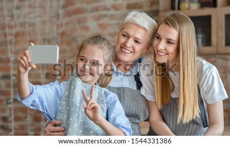 Cute little girl taking selfie with mom and granny in kitchen, girl showing victory sign, copy space