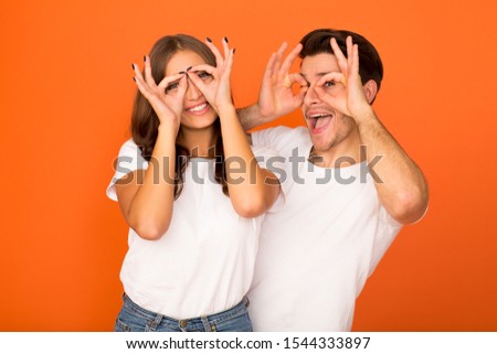 Crazy couple making funny glasses, holding fingers near eyes, orange studio background