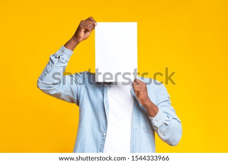 African american man covering his face with white blank paper, standing over yellow background in studio, copy space