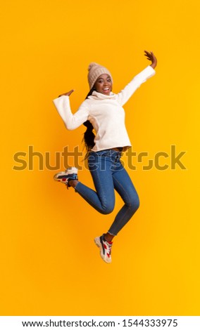 Lifeful afro girl in warm hat enjoying winter over yellow studio background, jumping up