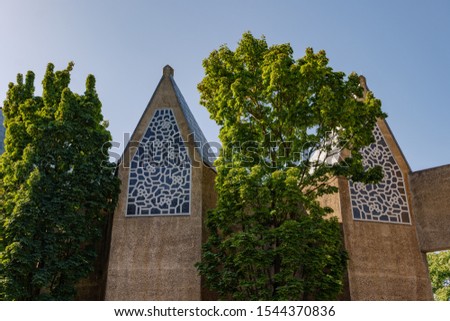 Outdoor sunny view of tree grow between  exterior sandstone concrete facade with abstract pattern ornament decorate on windows of brutalist architecture church in Cologne, Germany. 