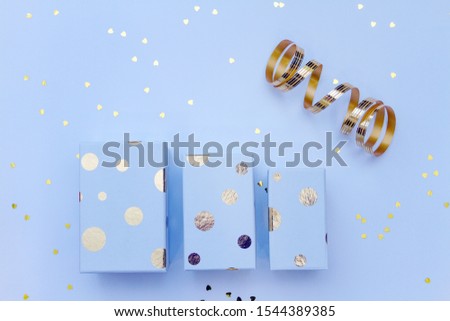 Three gift boxes with golden circles on a colored background with shiny hearts around. Blue holiday background.
