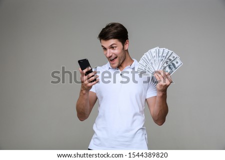 Image of emotional optimistic young man posing isolated over grey wall background holding money using smartphone.
