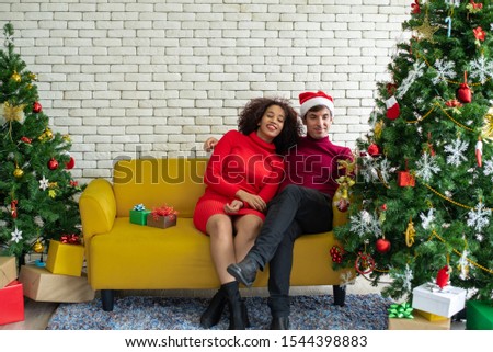 Happy young adult man hugging beautiful woman girlfriend and surprise her a Christmas present in gift box with Christmas tree decoration background. Christmas family celebration festive party concept.