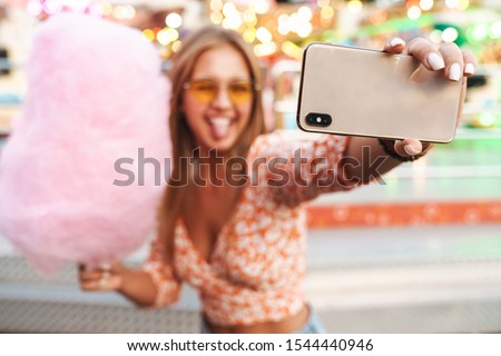 Photo of amazing pretty cute woman walking outdoors in amusement park eat candyfloss take selfie by mobile phone showing tongue.
