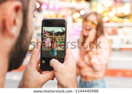 Image of a man take photo of his happy woman walking outdoors in amusement park by mobile phone.