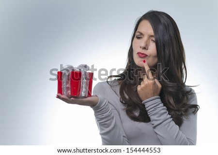 Beautiful young woman with long brunette hair standing holding an ornamental red gift on the palm of her hand looking at it with a puzzled thoughtful expression on a grey background with copyspace