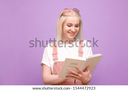 Happy blonde girl with a book in her hands on a purple background smiles and reads. Happy teenage girl in cute clothes reading a book and laughing, isolated on pink.