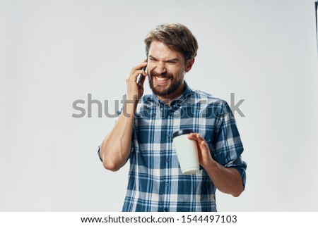 A handsome man in a plaid shirt is talking on the phone and a cup of coffee in his hand