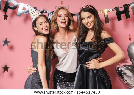 Image of three young party girls in festive outfits smiling and celebrating birthday isolated over pink background