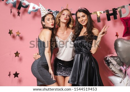 Image of three beautiful party girls in festive outfits smiling and celebrating birthday isolated over pink background