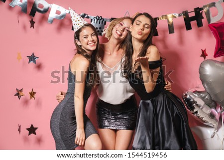 Image of three caucasian party girls in festive outfits smiling and celebrating birthday isolated over pink background