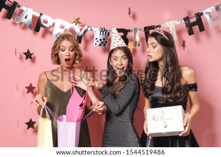 Image of adorable party girls in fancy dresses unpacking birthday gifts isolated over pink background