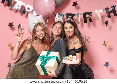 Image of pretty party girls in fancy dresses celebrating birthday with gift box and sweet cake isolated over pink background