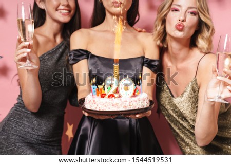 Image of attractive party girls in fancy dresses holding birthday cake and champagne glasses isolated over pink background