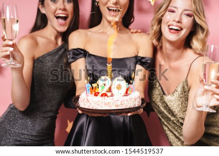 Image of gorgeous party girls in fancy dresses holding birthday cake and champagne glasses isolated over pink background