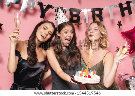 Image of cute party girls in fancy dresses holding birthday cake and champagne glasses isolated over pink background