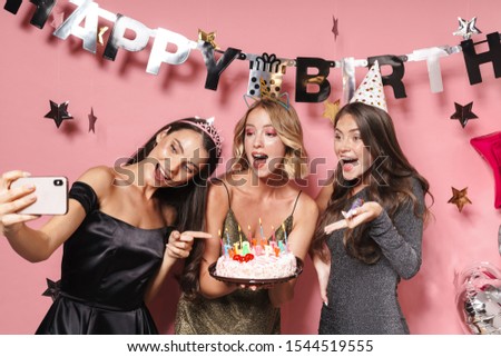 Image of delighted party girls taking selfie photo on smartphone and holding birthday cake with candles isolated over pink background