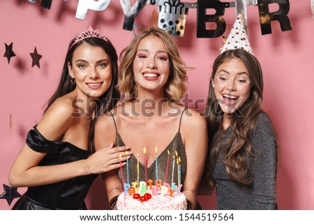 Image of adorable party girls in fancy dresses holding birthday cake with candles isolated over pink background