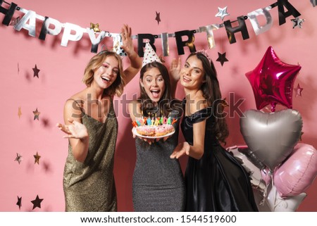 Image of beautiful party girls in festive dresses holding birthday cake with candles isolated over pink background