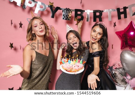 Image of joyful party girls in festive dresses holding birthday cake with candles isolated over pink background