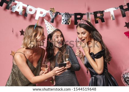 Image of cheerful party girls in festive dresses looking at smartphone while celebrating birthday isolated over pink background