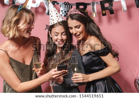 Image of adorable party girls in festive dresses looking at smartphone while celebrating birthday isolated over pink background