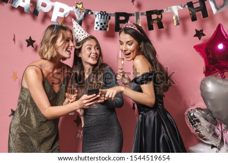 Image of alluring party girls in festive dresses looking at smartphone while celebrating birthday isolated over pink background