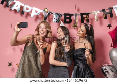 Image of young party girls in festive dresses taking selfie photo on smartphone while celebrating birthday isolated over pink background