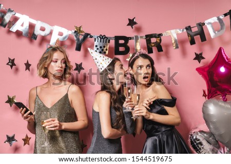 Image of three surprised party girls in festive dresses holding smartphone while celebrating birthday isolated over pink background