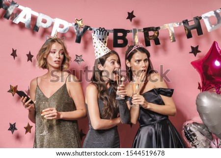 Image of three caucasian party girls in festive dresses holding smartphone while celebrating birthday isolated over pink background