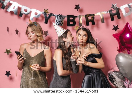 Image of three young party girls in festive dresses holding smartphone while celebrating birthday isolated over pink background