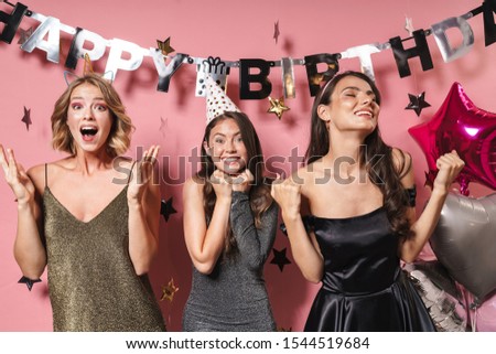 Image of three fashionable party girls in festive dresses smiling and celebrating birthday isolated over pink background